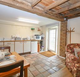 A kitchen area with cabinets a dining table set with dishes a floral armchair a coat rack and a door at Pear Tree Barn in Stalham