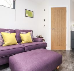 A living room with a purple sofa and ottoman yellow and floral cushions a wooden door and a black kitchen unit with a plant at Yew Tree Cabin in Ullswater