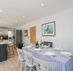 A dining area with a blue patterned tablecloth on a table set with white plates and glasses next to a kitchen with an island and three stools at Greystones in Upton St Leonards