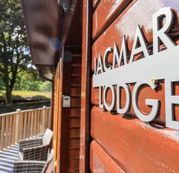 A wooden lodge exterior with a sign reading JacMar Lodge and outdoor chairs on a balcony surrounded by trees at JacMar Lodge 14 Applethwaite Limefitt Park near Windermere