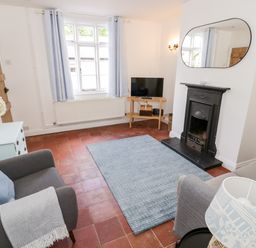 A living room with a fireplace a TV on a wooden stand two armchairs a blue rug and a window at Ross Cottage in Broadway