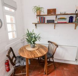 A small dining area with a round wooden table and two wooden chairs with cushions a potted plant on the table a white brick wall with wooden shelves holding books plants and decorative items a fire extinguisher near the window at Ross Cottage in Broadway
