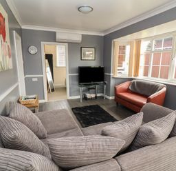 A living room with a large sofa and cushions a red armchair a television on a glass stand and windows at Swiss Cottage in Wilsthorpe near Bridlington
