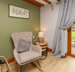 A corner of a room with a beige rocking chair a side table with a lamp and a basket of firewood next to a glass door with grey curtains at Pondarosa in Newbridge-On-Wye