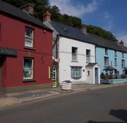 A street with red white and blue buildings including a shop and outdoor seating with umbrellas at Tan y Craig in Solva