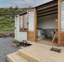 The open wooden door of a cabin showing a bed inside and outdoor seating on gravel in a rural area at Tan Y Castell Shepherds Hut in Llangollen