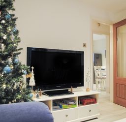 A living room with a decorated Christmas tree a flat screen TV on a white stand and a wooden door open to a dining area at 1 Camperdown Lodge in Salcombe