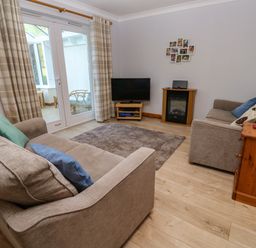 A living room with two beige sofas a television on a stand a small electric fireplace a wooden drawer unit and glass doors with curtains leading to a sunroom at Primrose Cottage Goldenbank near Falmouth