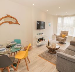 A living room with a round glass dining table with multicolored chairs a grey sofa patterned armchair a coffee table a wall-mounted TV and a dolphin-shaped mirror at Bay View in Criccieth