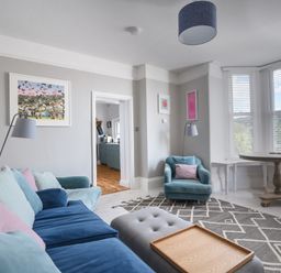 A living room with a blue sofa a blue armchair a gray ottoman wooden tray on ottoman round wooden table and large windows with blinds at Coastal View in Lyme Regis