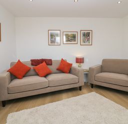 A living room with a beige sofa and armchair with orange cushions a small table with a lamp and framed pictures on walls at Dalegarth Hall Farm Cottage 1 Boot near Eskdale Green