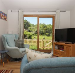 A living room with an armchair and a side table next to sliding glass doors with curtains a TV on a wooden cabinet and a sofa with patterned cushions at Adipit Stables in Buckland Brewer near Bideford