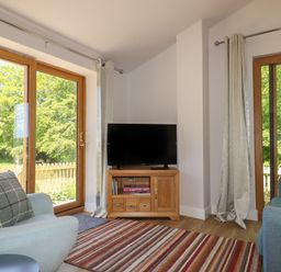 A living room with a TV on a wooden stand between two large windows at Adipit Stables Buckland Brewer near Bideford