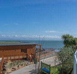 A wooden building with a green roof near the sea with a walkway and palm trees at E19 The Warren in Abersoch