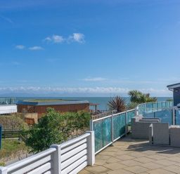 Outdoor terrace with wicker chairs glass railing overlooking sea and buildings at E19 The Warren in Abersoch