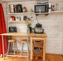 A small kitchen area with a wooden table and stools a microwave and kitchen appliances on shelves and a cart at Ty Bach Twt in Talybont-on-Usk