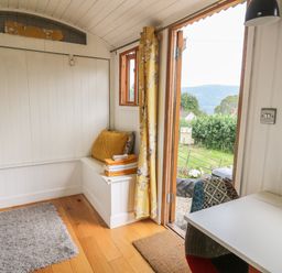 A small room with wooden floor a white bench with cushions and towels a table with a multicolored chair and an open door showing a green outdoor area at Ty Olwyn Bach in Talybont-on-Usk