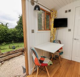 A small dining area with a white table, two patchwork chairs, a wall-mounted TV, a window with yellow curtains, and a door leading outside at Ty Olwyn Bach in Talybont-on-Usk