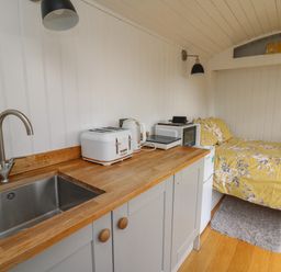 A kitchenette with a sink toaster kettle microwave and induction cooker next to a bed with yellow floral bedding at Ty Olwyn Bach in Talybont-on-Usk