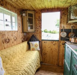 A small wooden kitchen with a yellow quilted couch two pillows a window a door a green cupboard and hanging pots at Ty Gwenyn in Talybont-On-Usk