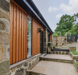 An exterior view of a stone and wood building with steps leading to a black door and a small garden space at The Bothy Riding Hills in Riding Mill near Corbridge