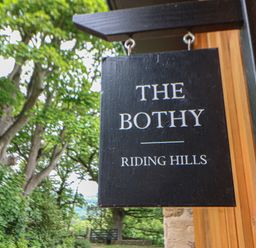 A black wooden sign with the text The Bothy Riding Hills hanging outside near trees and a wooden wall at The Bothy Riding Hills in Riding Mill near Corbridge