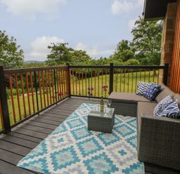 A wooden deck with a wicker sofa with blue patterned cushions a small table with a wine bottle and glasses and a blue patterned rug at The Bothy Riding Hills in Riding Mill near Corbridge