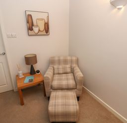 A corner with a plaid armchair and matching ottoman next to a wooden side table with a lamp and decorative items at Flat 1 Tenby