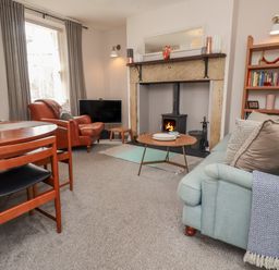 A living room with a blue sofa, wooden dining table, leather chair, burning wood stove, and bookshelf at White Feathers in Alnwick