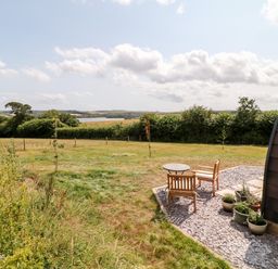 A grassy field with a small patio area with wooden chairs and a table beside a curved building at Oak in Elmgate near Saltash