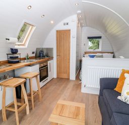 A compact kitchen with wooden stools a small oven and a sink next to a living area with a blue sofa and cushions at Oak Elmgate near Saltash
