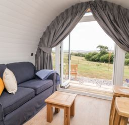 A small living area with a dark blue sofa with pillows a wooden coffee table and stools near open glass doors with curtains overlooking a grassy landscape at Oak in Elmgate near Saltash