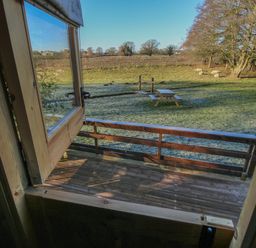 A wooden deck with a railing viewed through an open window overlooking a grassy yard with a picnic table and sheep at The Oak Hut in Coton near Whitchurch