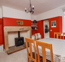 A dining room with a polka dot tablecloth on a table wooden chairs a black stove in a stone fireplace red walls and a window at Victoria House in Alnwick