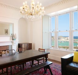 A dining room with a large wooden table, chairs, a chandelier, a fireplace, and a window overlooking a harbor at Waves End in St Ives