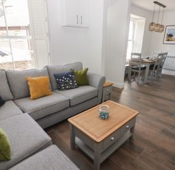 A living room with a grey sofa with cushions and a wooden coffee table next to a dining area with a wooden table and chairs at Gwynfryn in Pen-Clawdd