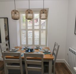 A dining room with a wooden table set for six under three pendant lights and a window with shutters at Gwynfryn in Pen-Clawdd
