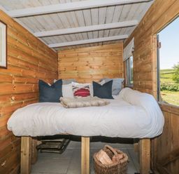 A wooden cabin interior with a bed elevated on wooden legs with pillows and a basket of firewood underneath at The Snug in Dulverton