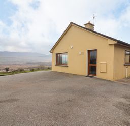 A single-story house with yellow walls and several windows on a paved area with hills in the background at Gortnamona in Coolnaharragill Lower near Glenbeigh County Kerry