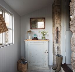 A hallway with a white cabinet topped with a lantern, flowers, and binoculars next to stone walls and boots by the door at Butterwell Cottage at Collihole in Chagford