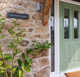 A green front door with glass panels next to a stone wall with a plant and a sign that reads Chynoweth House at Chynoweth House in Cubert