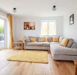 A living room with a gray sectional sofa with yellow cushions a yellow rug wooden tables a lamp and framed pictures at Chynoweth House in Cubert
