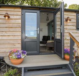 An open door to a wooden cabin with potted flowers on the porch at Shepherds Stone in Llanfairynghornwy near Llanfaethlu
