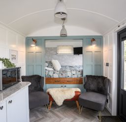 A small living area with two chairs and a bench in front of a built-in bed alcove with a patterned bedspread and a small mounted TV at Shepherd's Stone in Llanfairynghornwy near Llanfaethlu