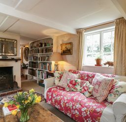 A living room with a sofa covered in a red floral throw pillows a coffee table with a vase of flowers a window a fireplace and a bookshelf at Chalkpit Cottage in Blewbury