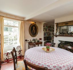 A dining room with a round table covered with a tablecloth and chairs around it and a fireplace with a large mirror above and a bookshelf in the corner at Chalkpit Cottage in Blewbury