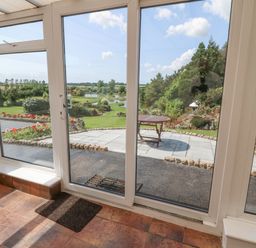 A sunroom with tiled floor and glass doors leading to a garden with patio table and landscaped greenery at Pen Bonc  Llanddaniel Near Gaerwen