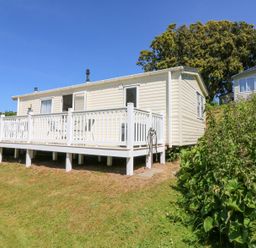 An exterior view of a mobile home with a white deck and railing on a grassy slope with shrubs and trees at Horizon in Combe Martin