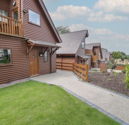 A walkway leading to brown log cabins with green lawn and garden at Classic Lodge 16 in Dyserth