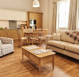 A living room with a sofa a chair a wooden coffee table with newspapers and an open kitchen with a dining table and chairs at The Stalls in Humshaugh